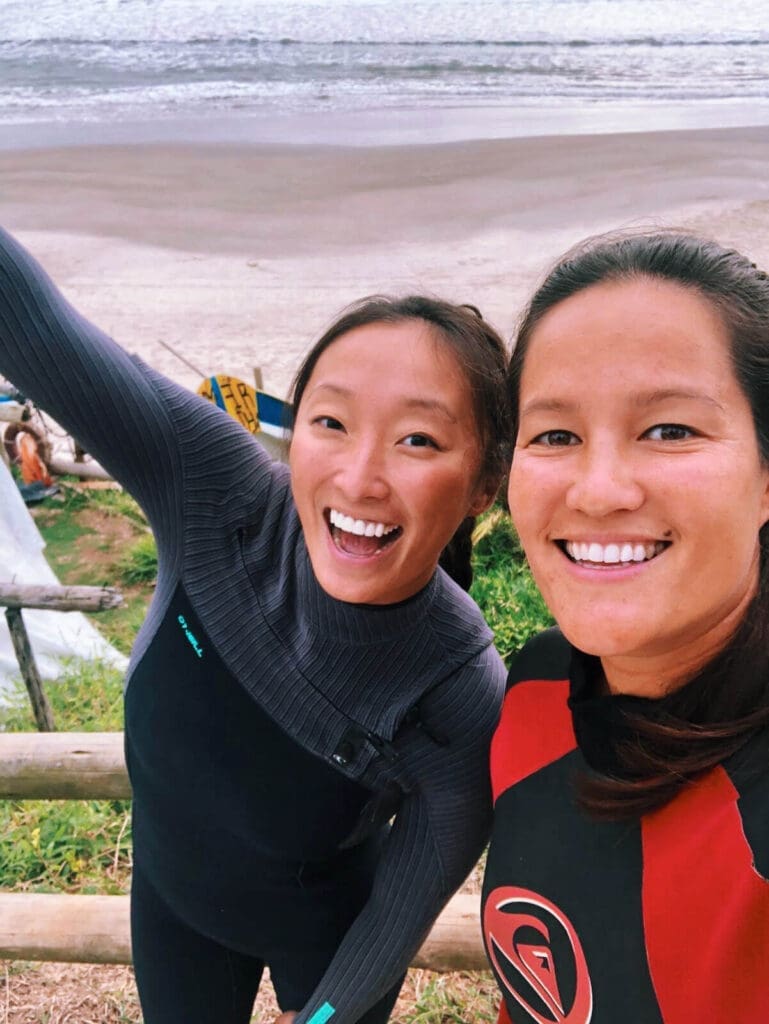 Two girls in wetsuits smiling at the camera with the beach in the back