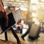 A traveller walking through an airport terminal, holding a passport, a boarding pass, and a leather duffel bag.